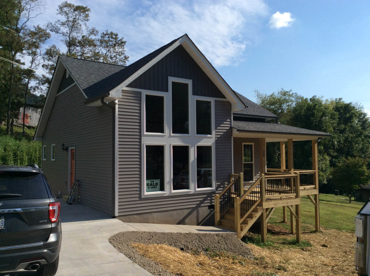 Two-story house with gray siding, covered front porch featuring black railings, parked car in driveway, large windows, and landscaped yard with trees under a cloudy sky