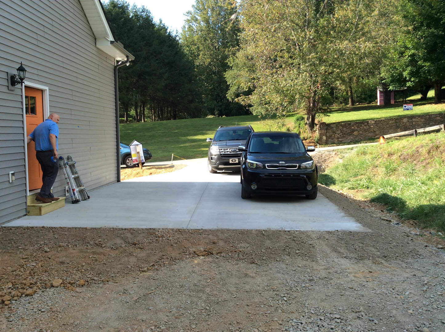 Black sedan parked on driveway beside a man in blue shirt, with green-leaved tree and grass in background