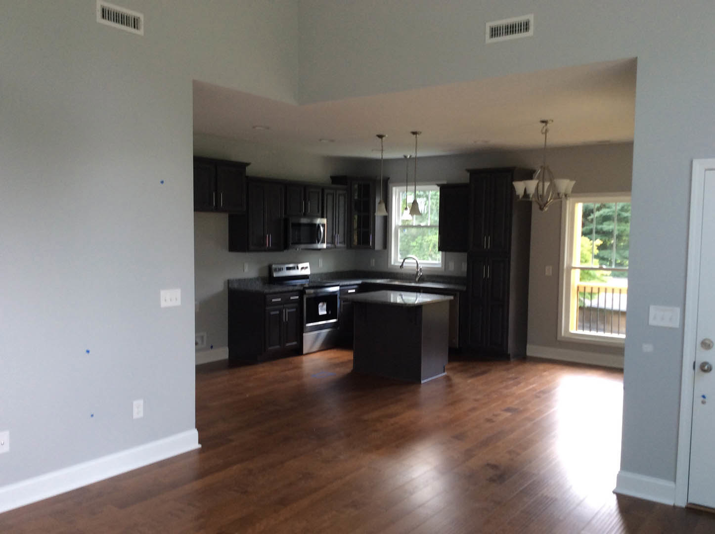 Kitchen with hardwood flooring, white cabinetry, stainless steel appliances, black glass cooktop, wall vent, and window overlooking trees with a railing.