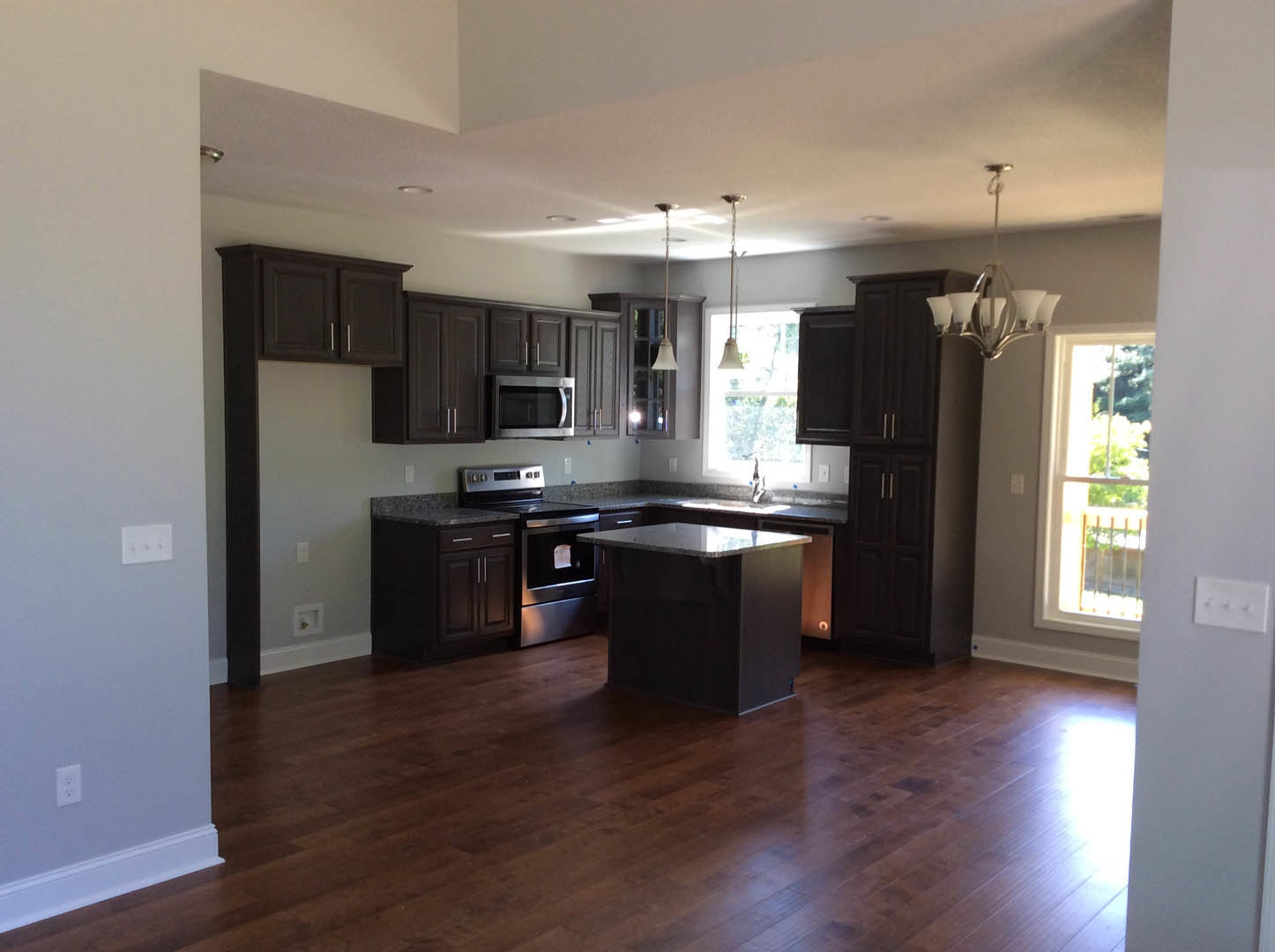 Kitchen with dark wood cabinets, marble-topped island, wood flooring, stainless steel microwave, stove with paper taped to oven door, white light switch on wall, overhead lighting