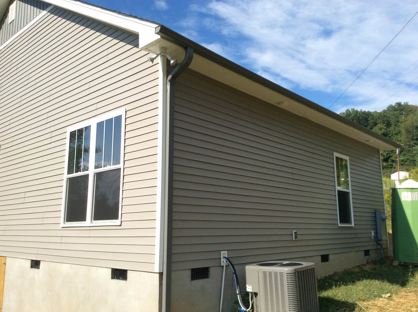White siding exterior of a single-story home with a large window and wall-mounted heater vent, surrounded by grass and partly cloudy sky.