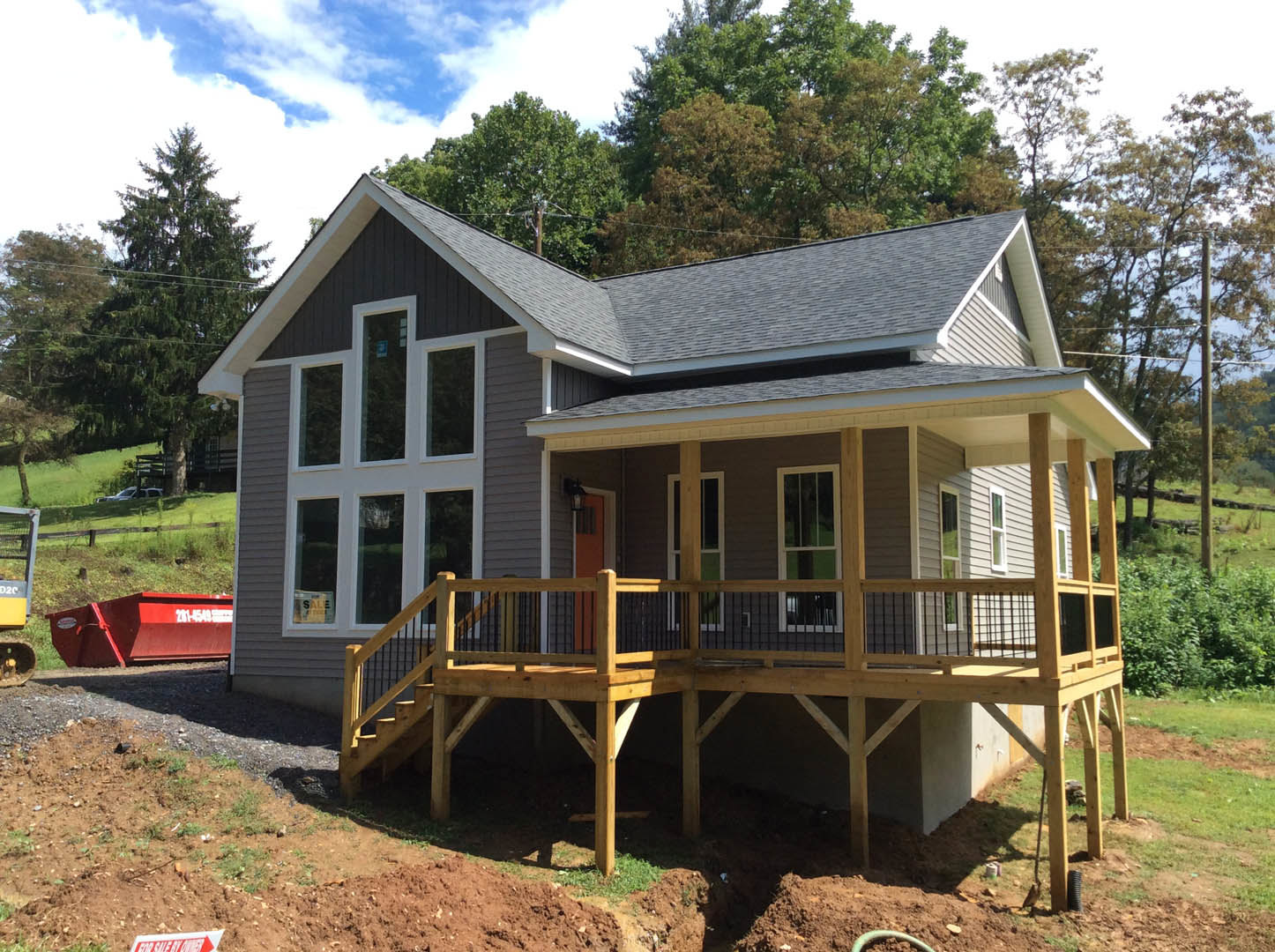 Two-story house with gray siding, white trim, wooden deck with railings, covered front porch, large windows, red dumpster in driveway, blue sky and green trees in background