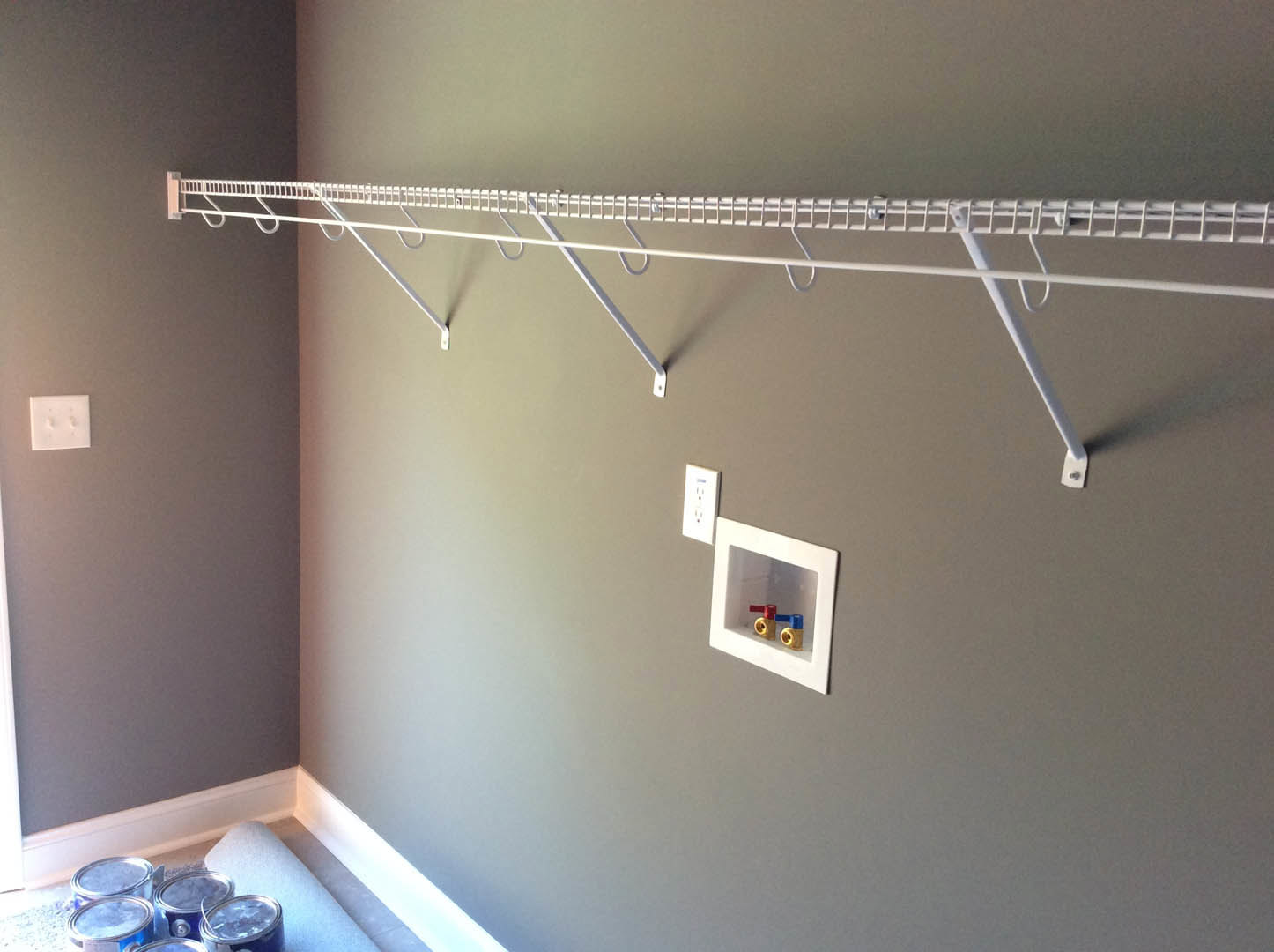 White metal rack mounted on a wall with colorful pipes, paint cans, and a power outlet nearby.