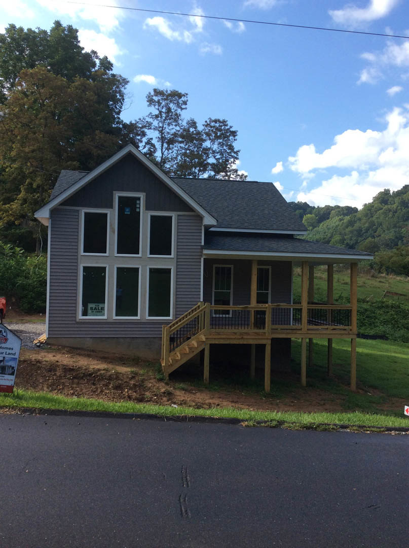 Two-story house with white siding, covered front porch with wooden railings, concrete driveway, large windows, and landscaped yard with trees.