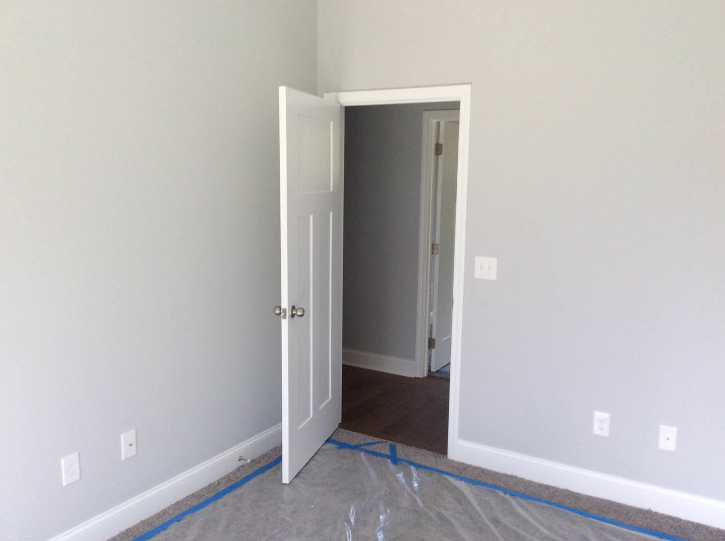 White door with silver knobs partially open in a room, plastic wrap covering the floor, white wall outlet and two-switch light switch on smooth plaster wall