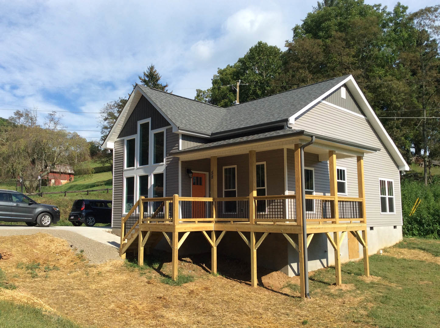 Two-story house with white-framed windows, wooden porch and railing, black car parked on grass behind the home, surrounded by trees and cloudy sky
