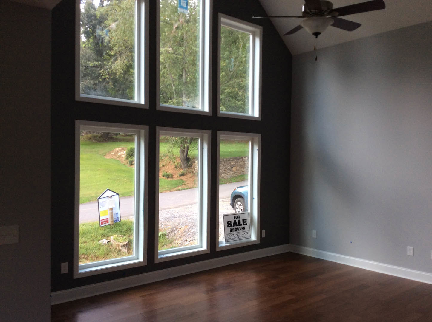 Spacious room featuring multiple tall windows with white trim, wood flooring, ceiling fan with light fixture, and a white sign with black text visible near one window.