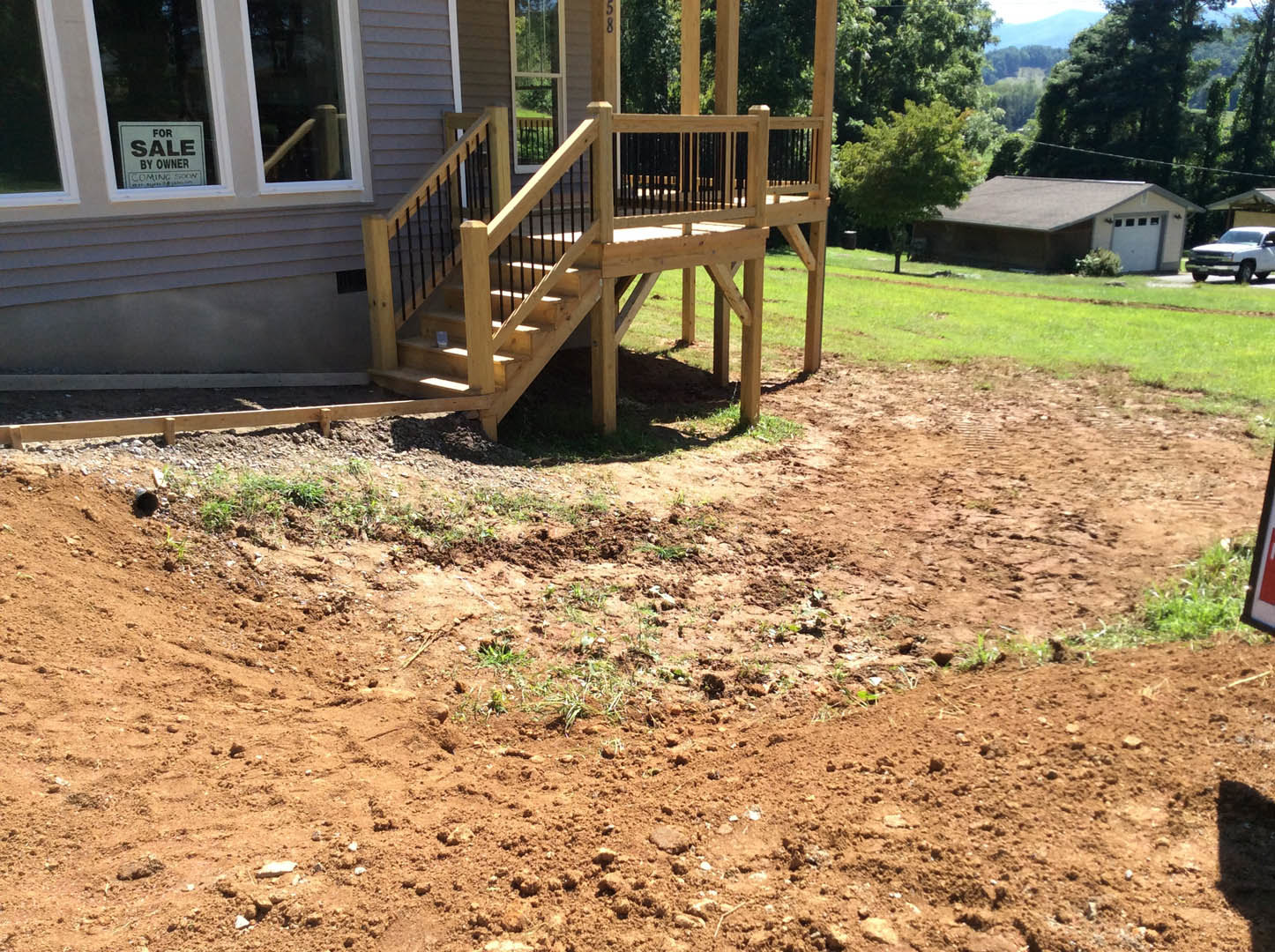 Two-story house with covered porch and wooden stairs, white siding, wooden deck with railings, dirt path leading to entry, grassy yard, mature trees, small outbuilding with gabled
