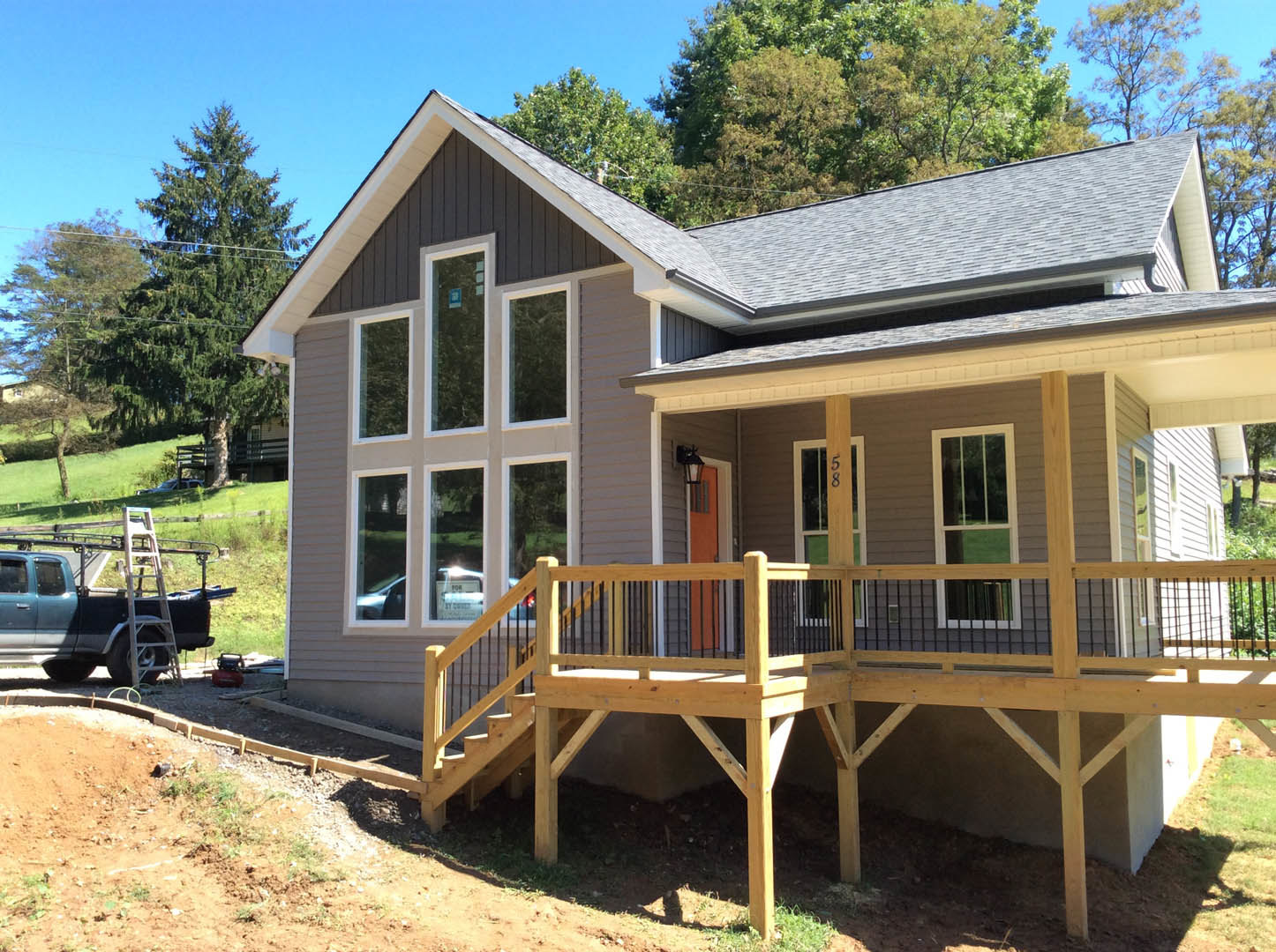 Wooden deck with railing and covered porch on a residential home, white-framed windows, blue sign in window, ladder secured to a parked truck, mature trees in background.
