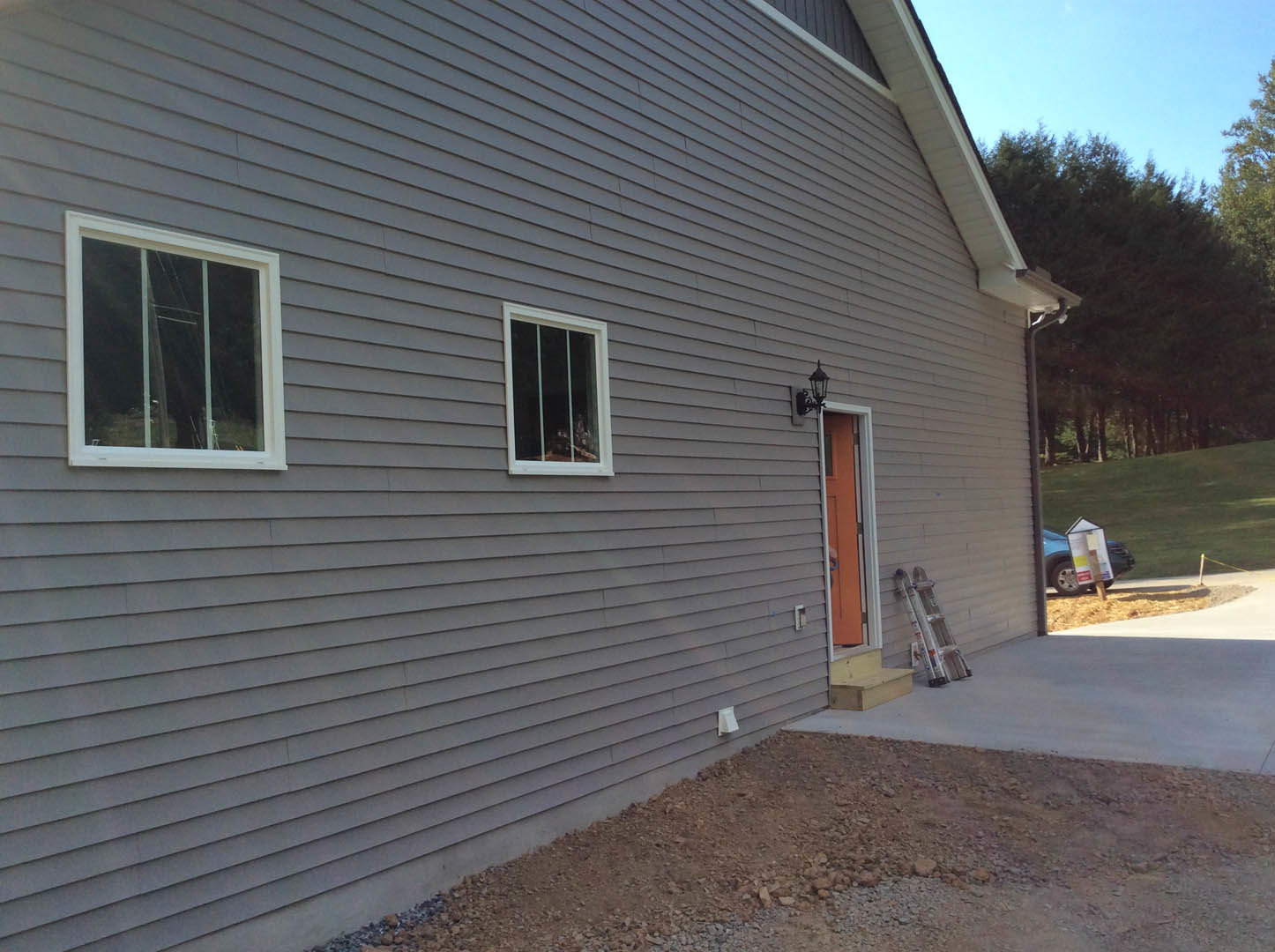 Grey two-story house with white-framed windows, concrete driveway, ladder leaning against exterior wall, trees in background, person visible through window