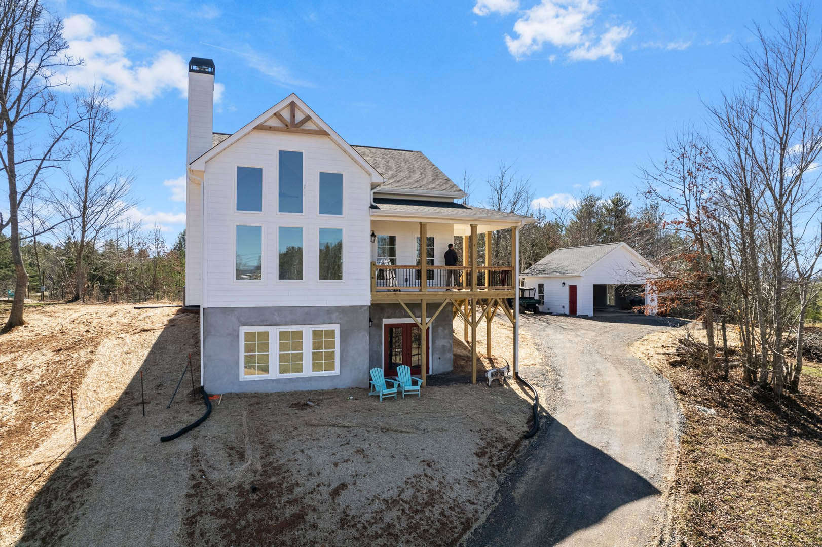White house with multiple windows, red front door, covered porch with blue chair, concrete driveway, green lawn, trees, and blue sky with scattered clouds