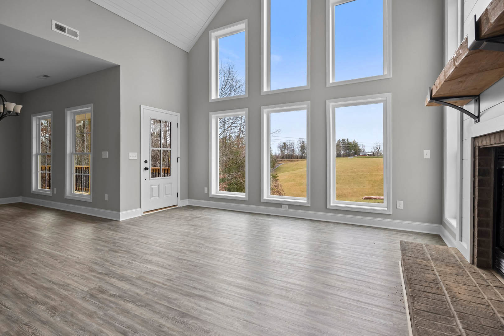 Spacious room with wide wood flooring, multiple large windows, white door with glass panel, brick step, and view of trees and open field through windows