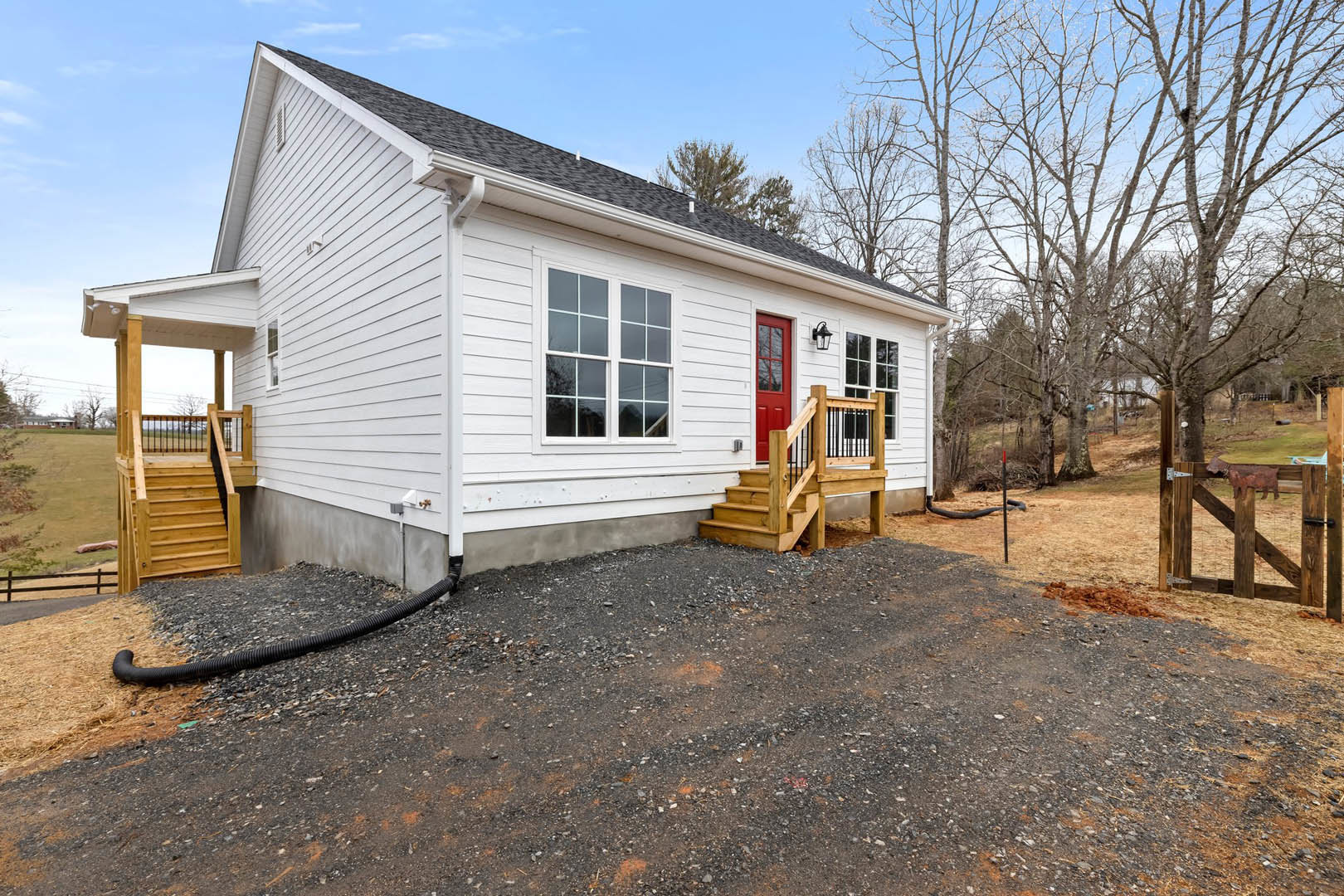 White siding house with a red front door, gravel driveway, wooden exterior stairs, black pipe along the ground, and trees in the background