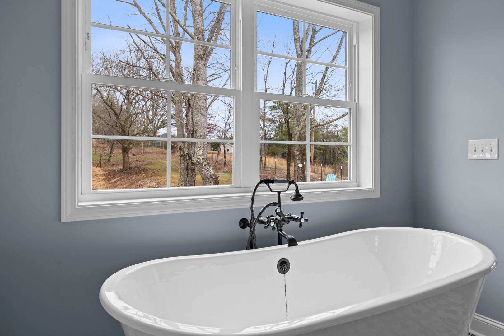 Freestanding white bathtub with black faucet and hose beside large windows overlooking leafy trees, white walls, and bathroom accessories on nearby countertop