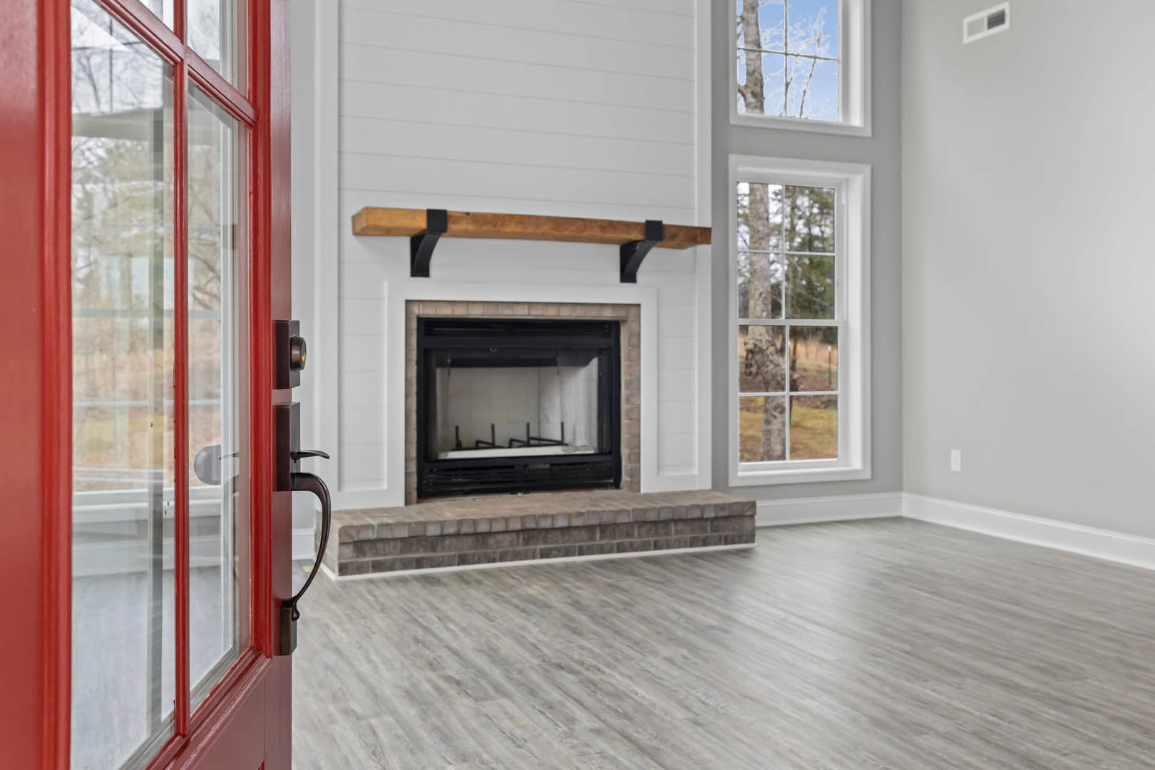 Living room with brick fireplace, glass window, red door, wood laminate flooring, staircase, and large window overlooking trees.