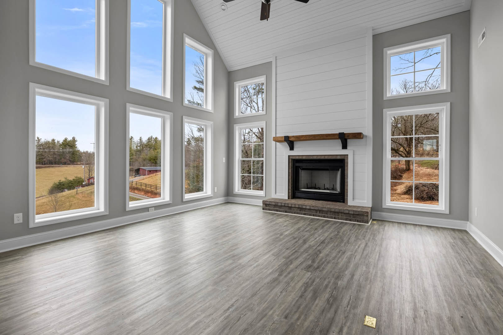 Living room with black-framed fireplace set in a brick accent wall, hardwood floors, large windows overlooking trees, and a gold square decor piece on a wooden surface.