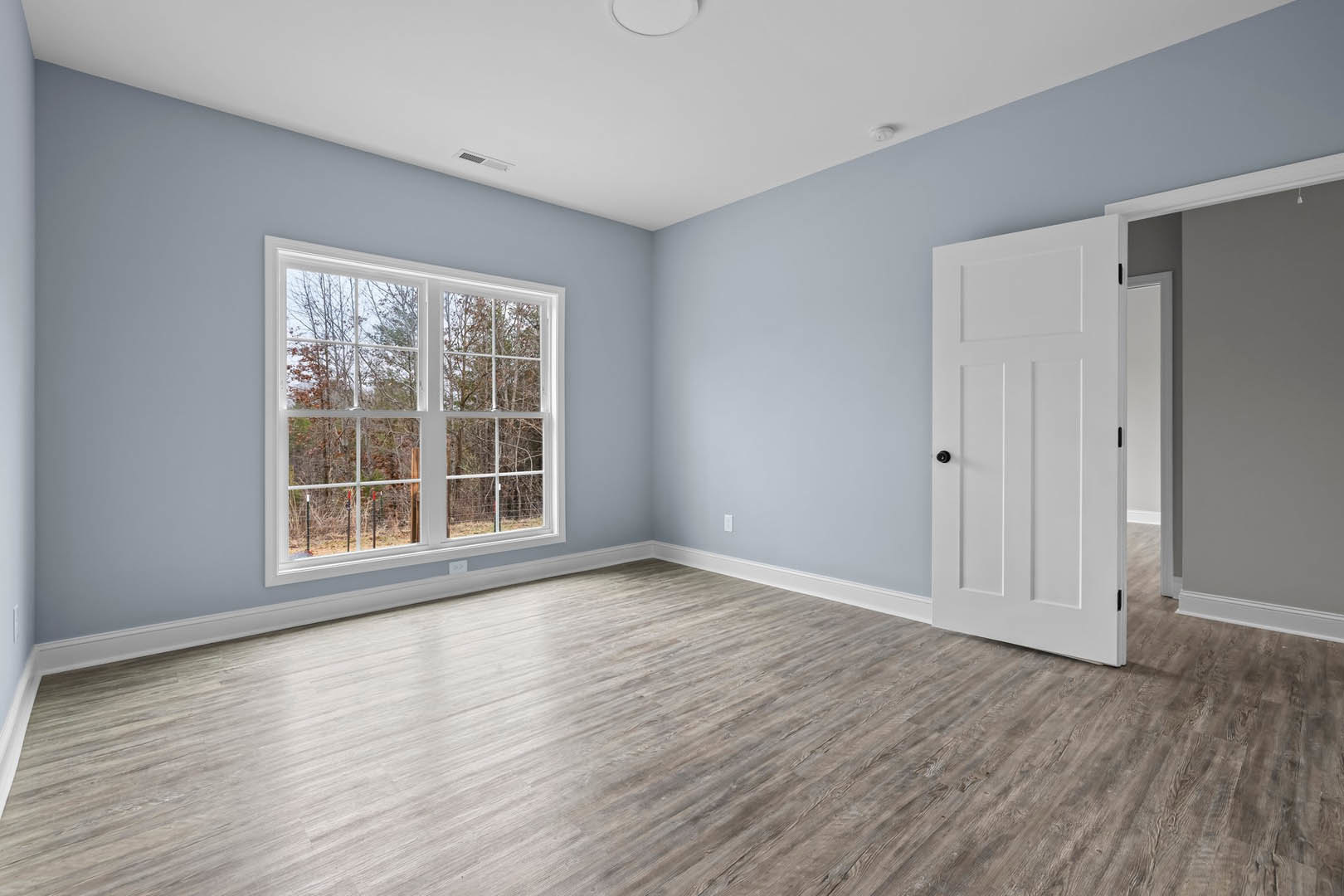 White-walled room with wood flooring, white door featuring black knobs, large window framed in white, trees visible outside, smooth white ceiling