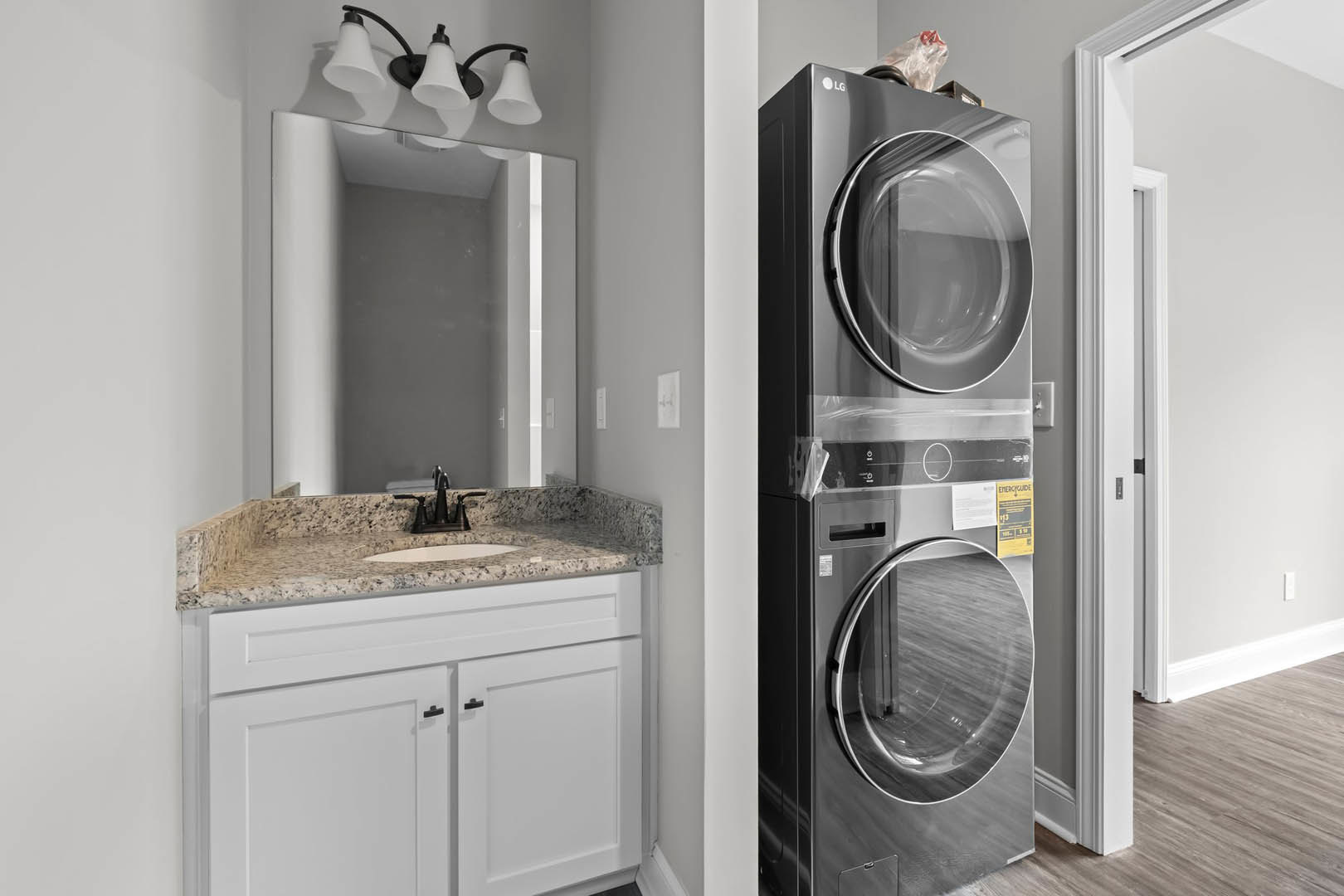 Front-loading washer and dryer beside white bathroom sink with chrome faucet, light fixture above, cabinetry and doorway visible