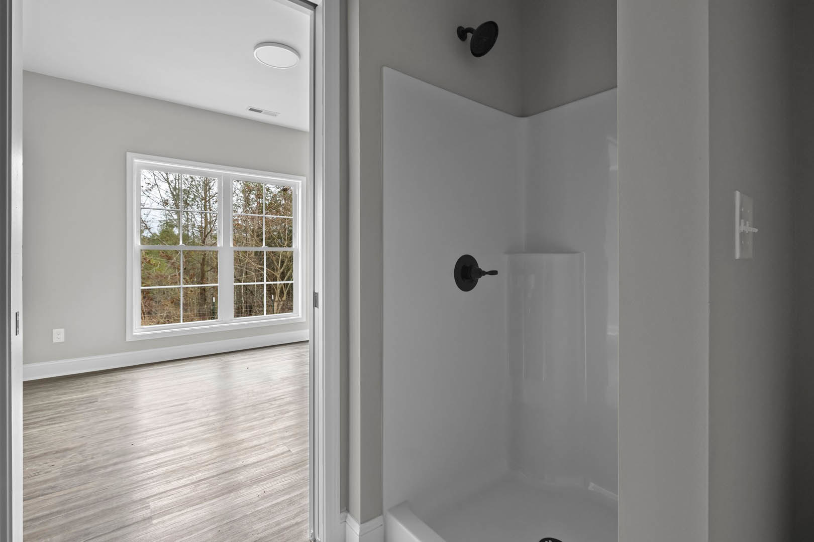 White shower enclosure with black handle and shower head, wood flooring with white trim, window overlooking trees, close-up of a round black knob on the door