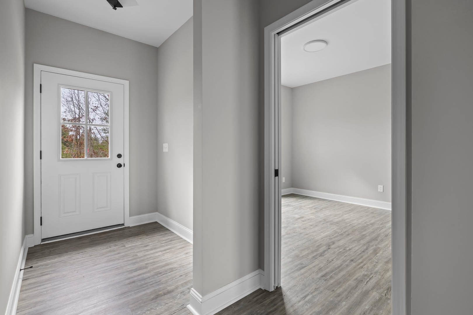 Hallway with light wood laminate flooring, white walls, white ceiling with round flush light, white door featuring a window, open doorway leading to another room, window showing
