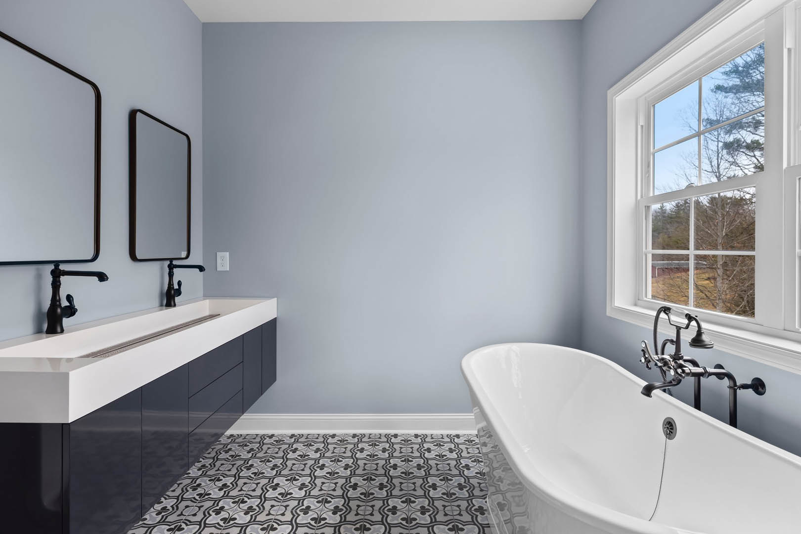 White freestanding bathtub with matte black faucet beside a window overlooking trees and blue sky, white sink with black handles on tiled wall, phone resting near tub