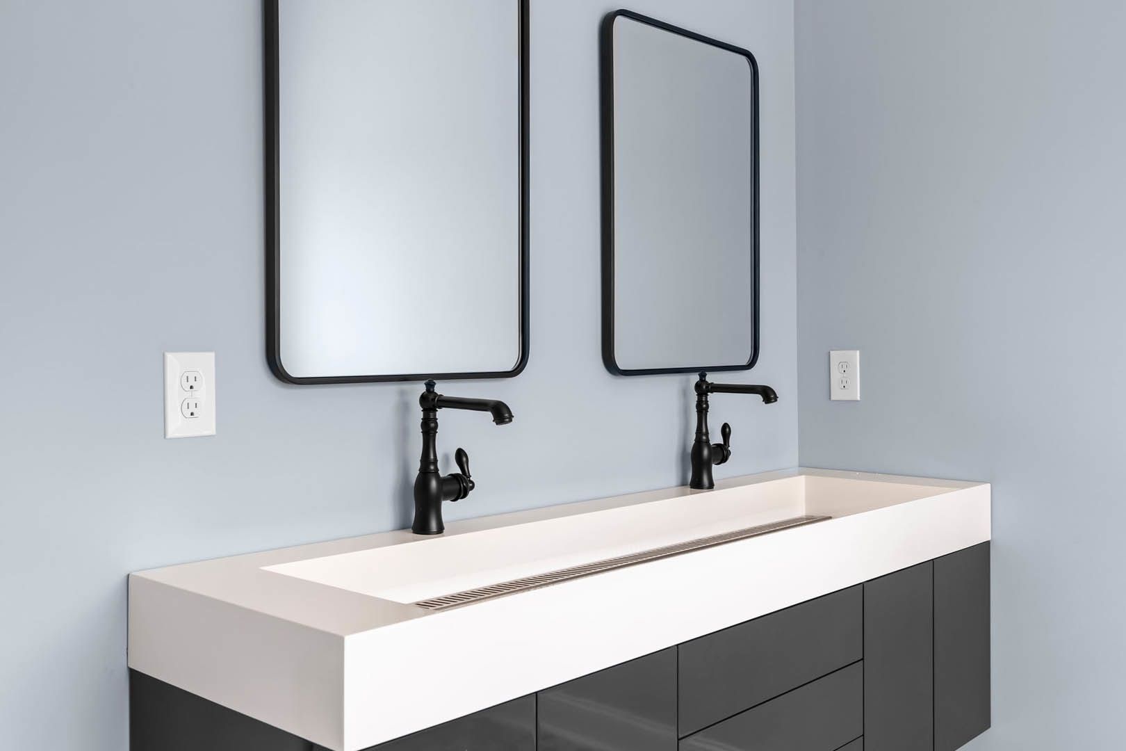 White bathroom sink with black faucet and handles, two rectangular mirrors with black frames mounted on a white wall, electrical outlet visible near the sink.