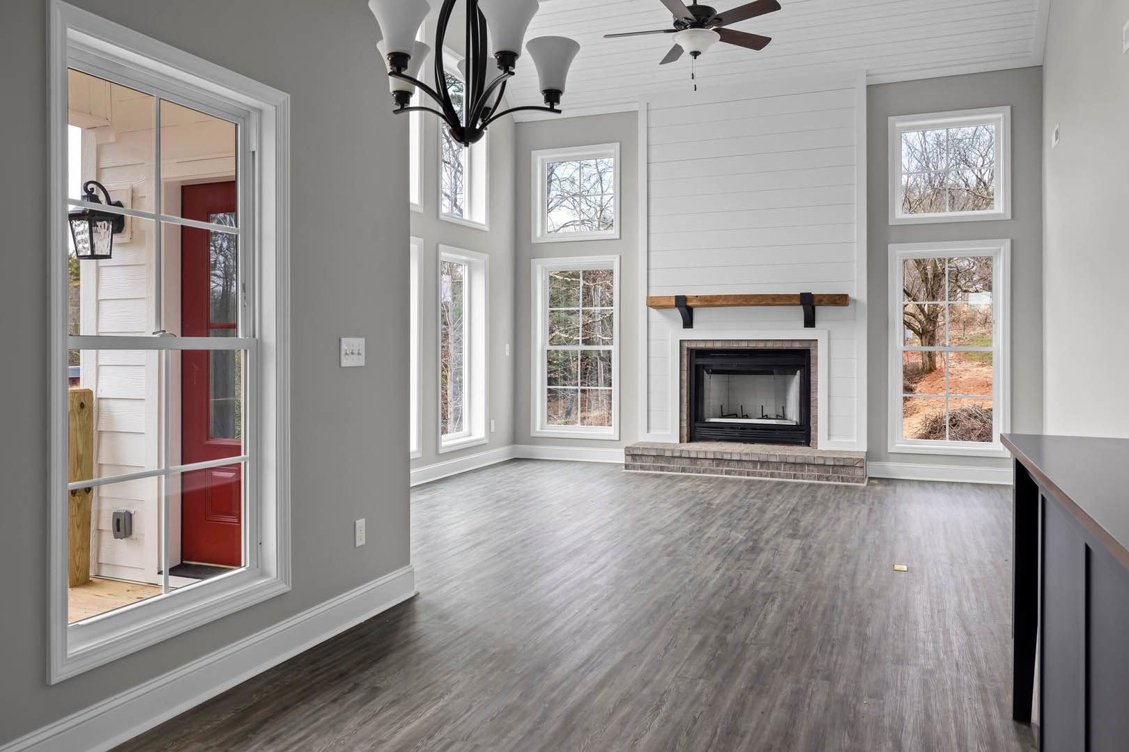 Living room with black-framed fireplace, ceiling fan, hardwood floors, chandelier, large windows showing trees, and light cabinetry