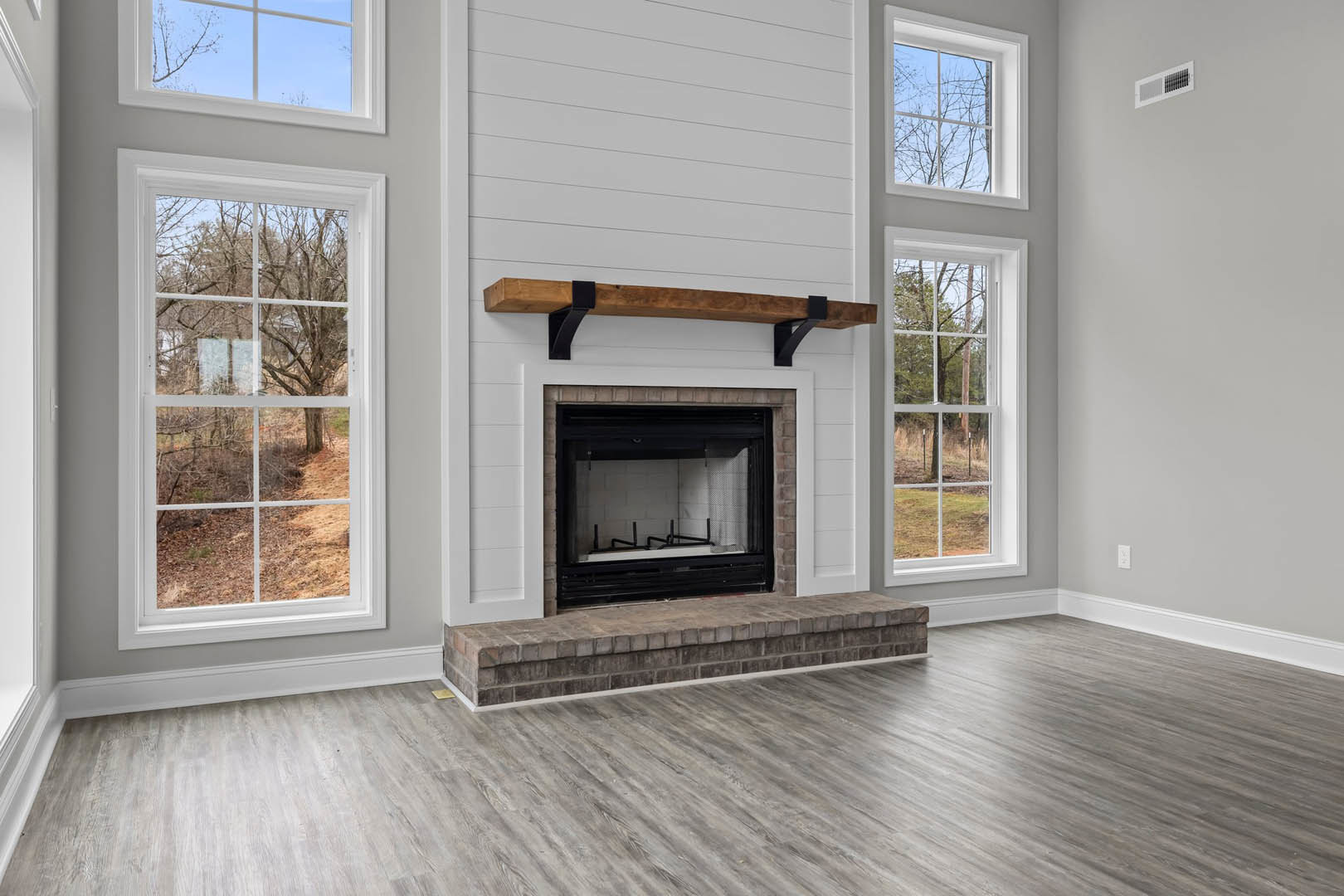 Stone fireplace with black frame set in white wall, wood flooring, exposed wooden ceiling beam, large windows showing trees and bare ground outside
