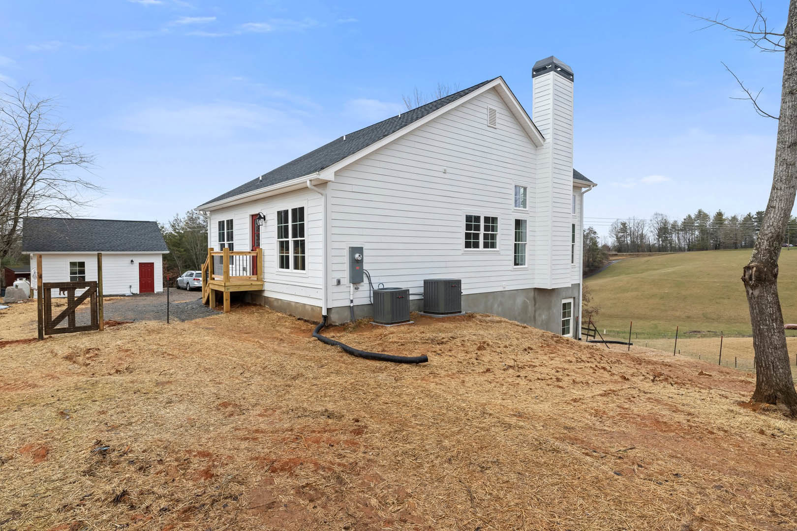 White house with wooden deck featuring metal railing, spacious grassy yard, wooden gate with a dog, porch, fence, window, and black pipe lying on the ground.