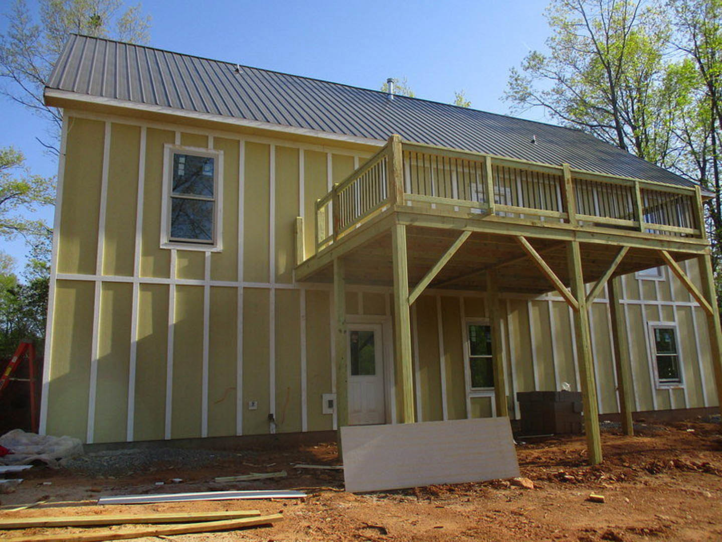 Partially built home with exposed wooden deck, white siding, porch area, large windows, and construction materials scattered on bare ground