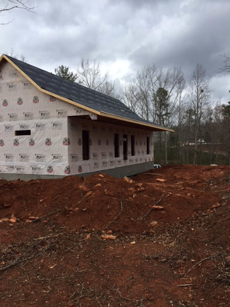 Partially built house with exposed roof framing, surrounded by dirt piles and mature trees under a cloudy sky