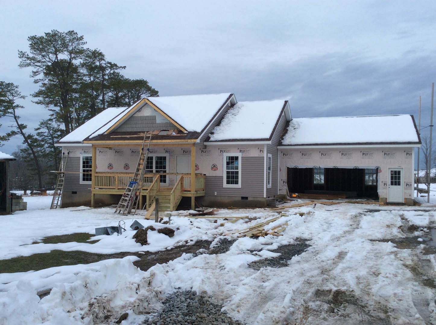 Partially built house with snow covering the roof, white door with window, multi-pane windows, pile of snow and rocks in front, ladder leaning against exterior wall, bare trees and