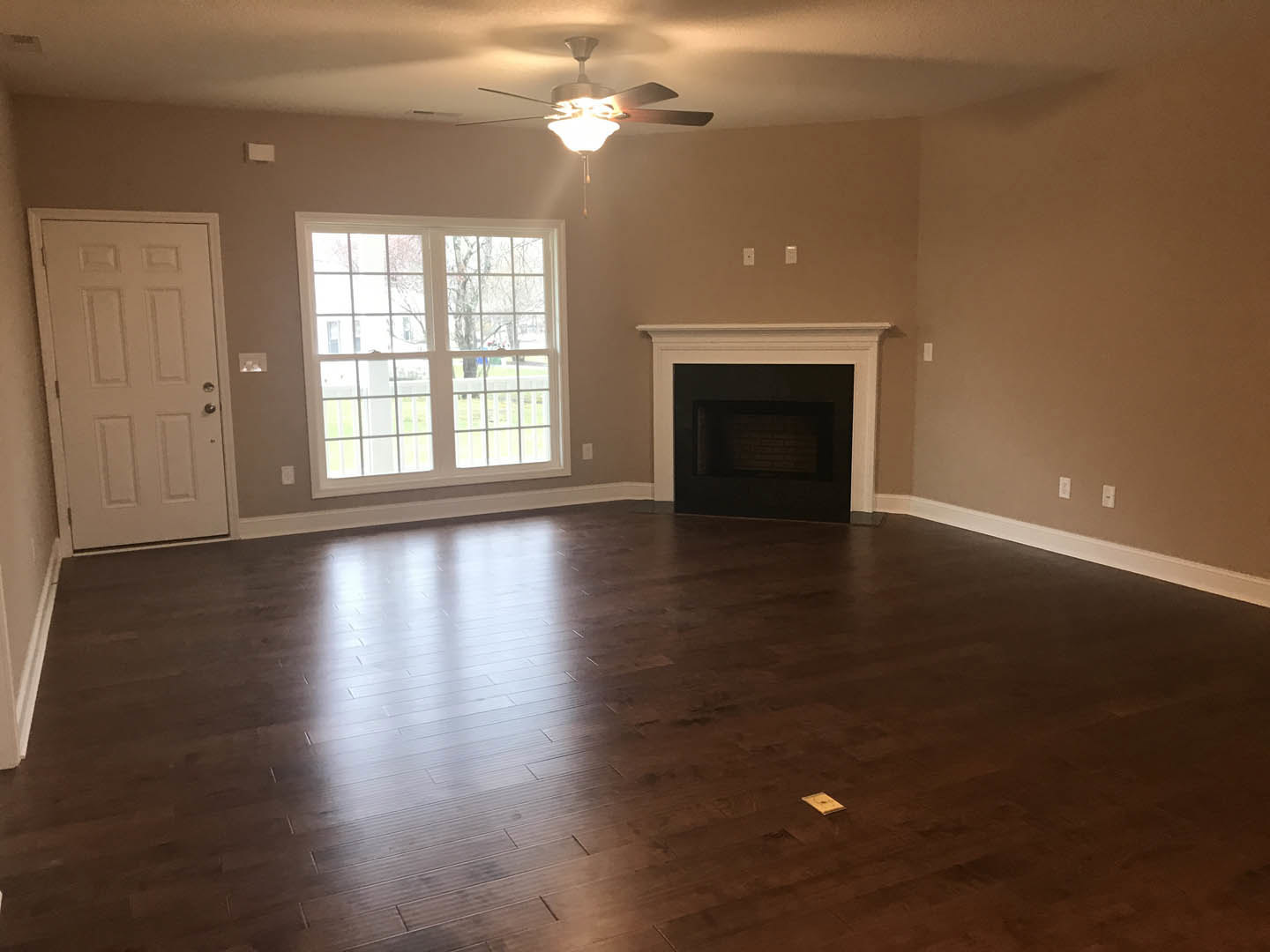 Living room featuring multi-pane window, hardwood flooring, white door with silver handle, brick-accented fireplace with black surround, and ceiling fan with illuminated light.