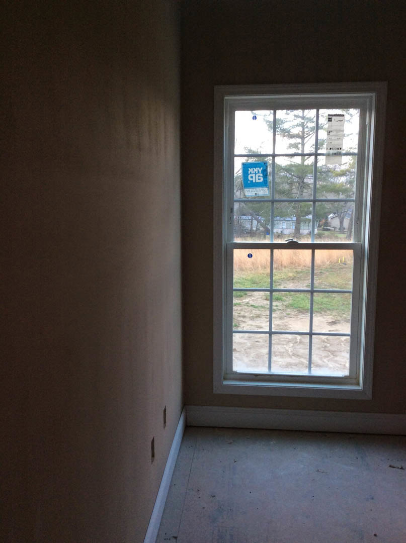 Large window with blue sign and white text, set in a grey wall, overlooking grassy field; interior room features light flooring and minimal decor.