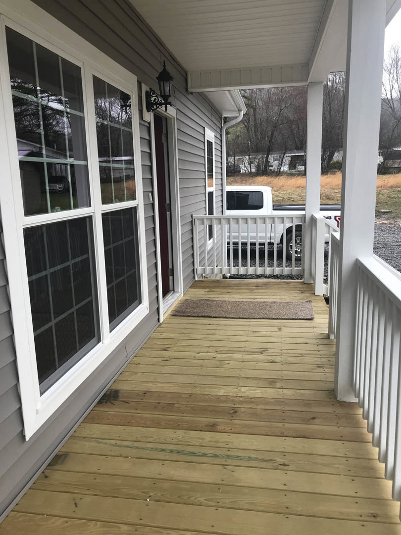 Front porch with white railing, wooden deck, white-framed window, entry door, and welcome mat; white fence and parked car visible in background