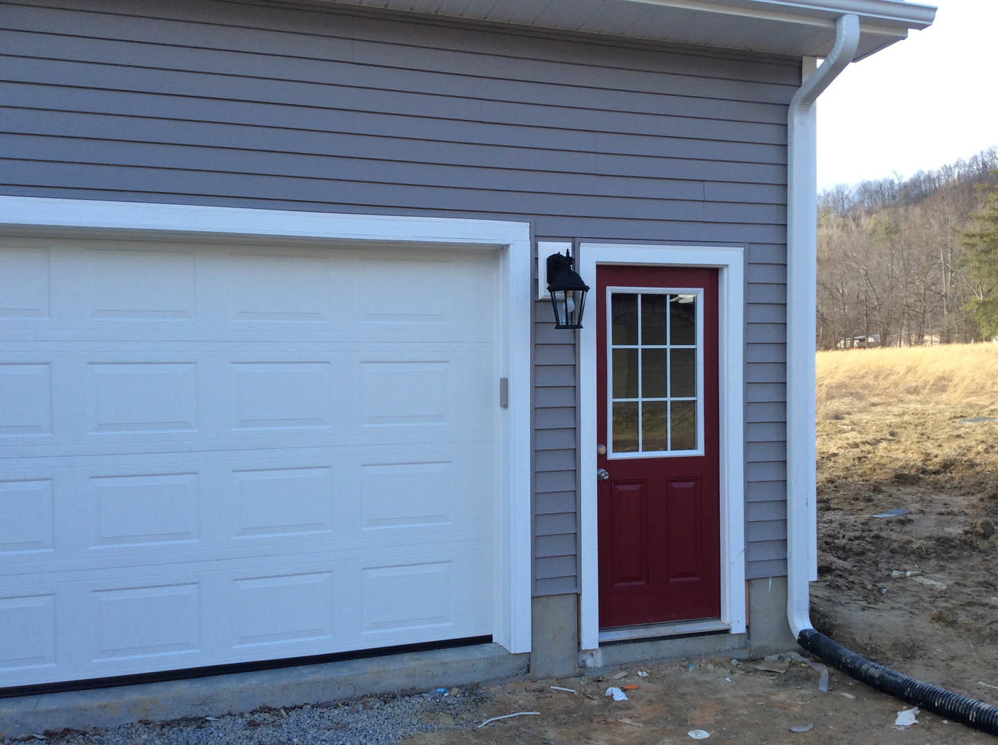 White garage door set in gray siding, adjacent to a red entry door with white trim, single window with white frame, outdoor light fixture above, trees visible on hillside in