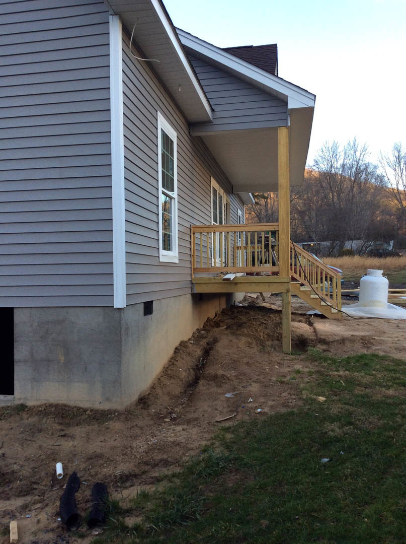 Modern home with light siding, elevated wooden deck, staircase leading to grassy yard, concrete retaining wall, scattered outdoor utility containers, and surrounding trees.