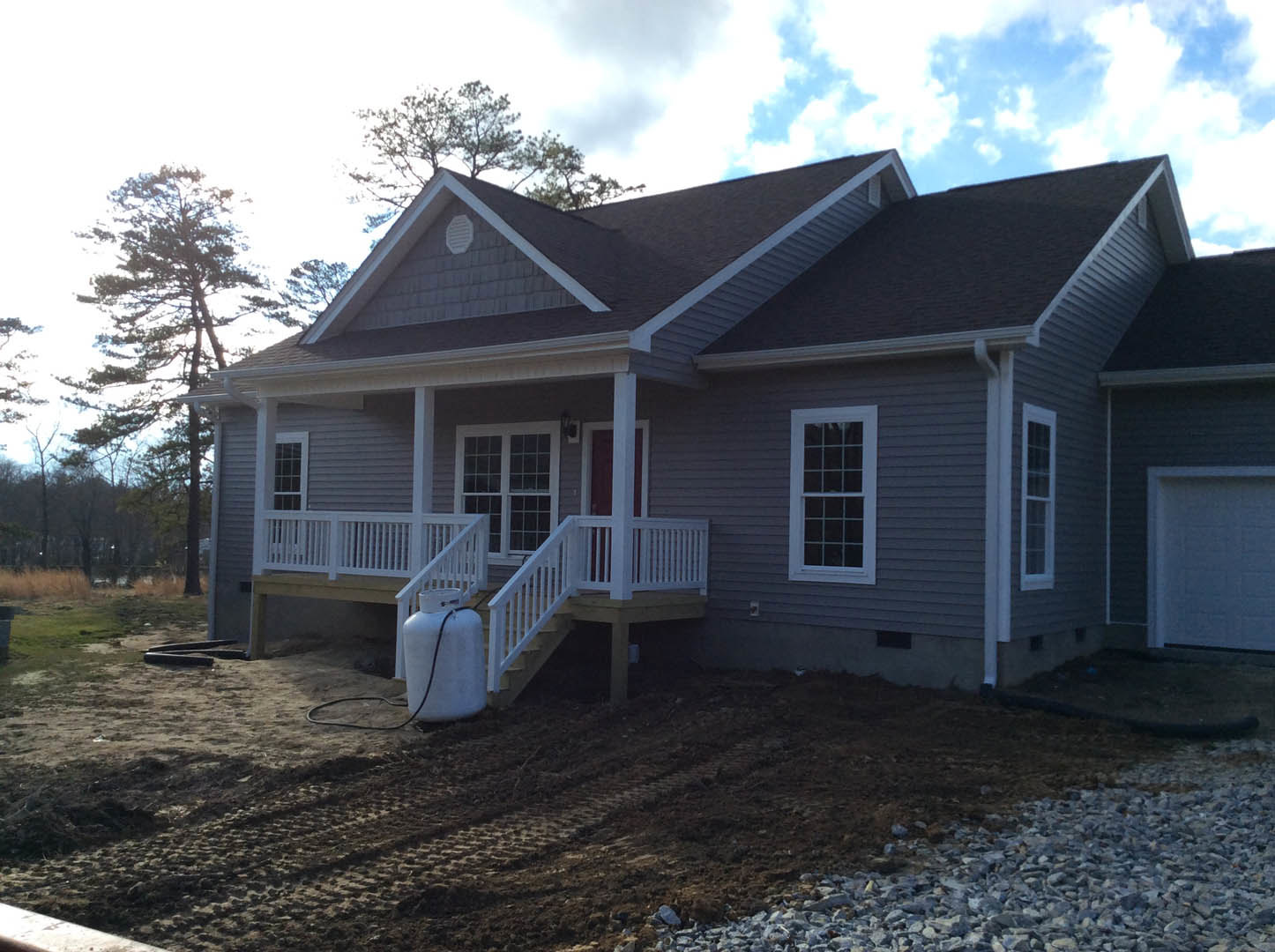 White deck with railing, porch stairs, blue exterior wall with white brick accents, multi-pane window, white tank with black cord on dirt patch, cloudy sky above