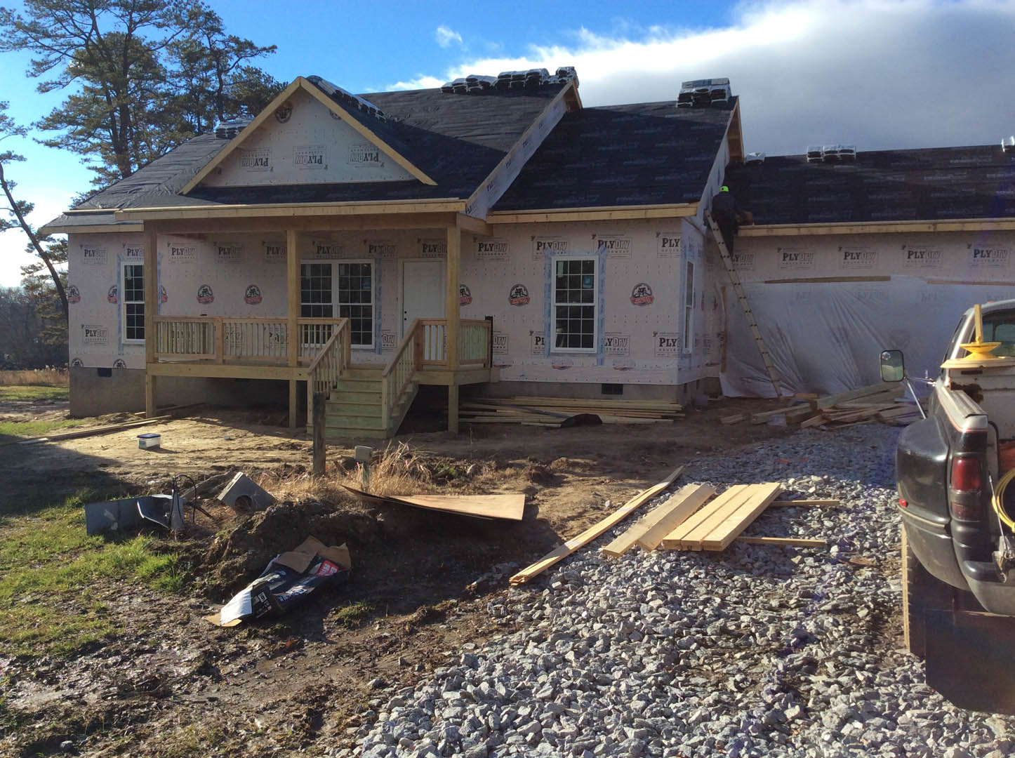 Two-story house under construction with exposed framing, gravel driveway, wooden deck and stairs, surrounded by mature trees and cloudy sky