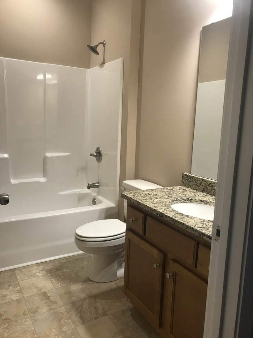 Bathroom with white toilet, white bathtub featuring silver fixtures, wall-mounted shower head, stone accent wall, white countertop, and partial view of a door.