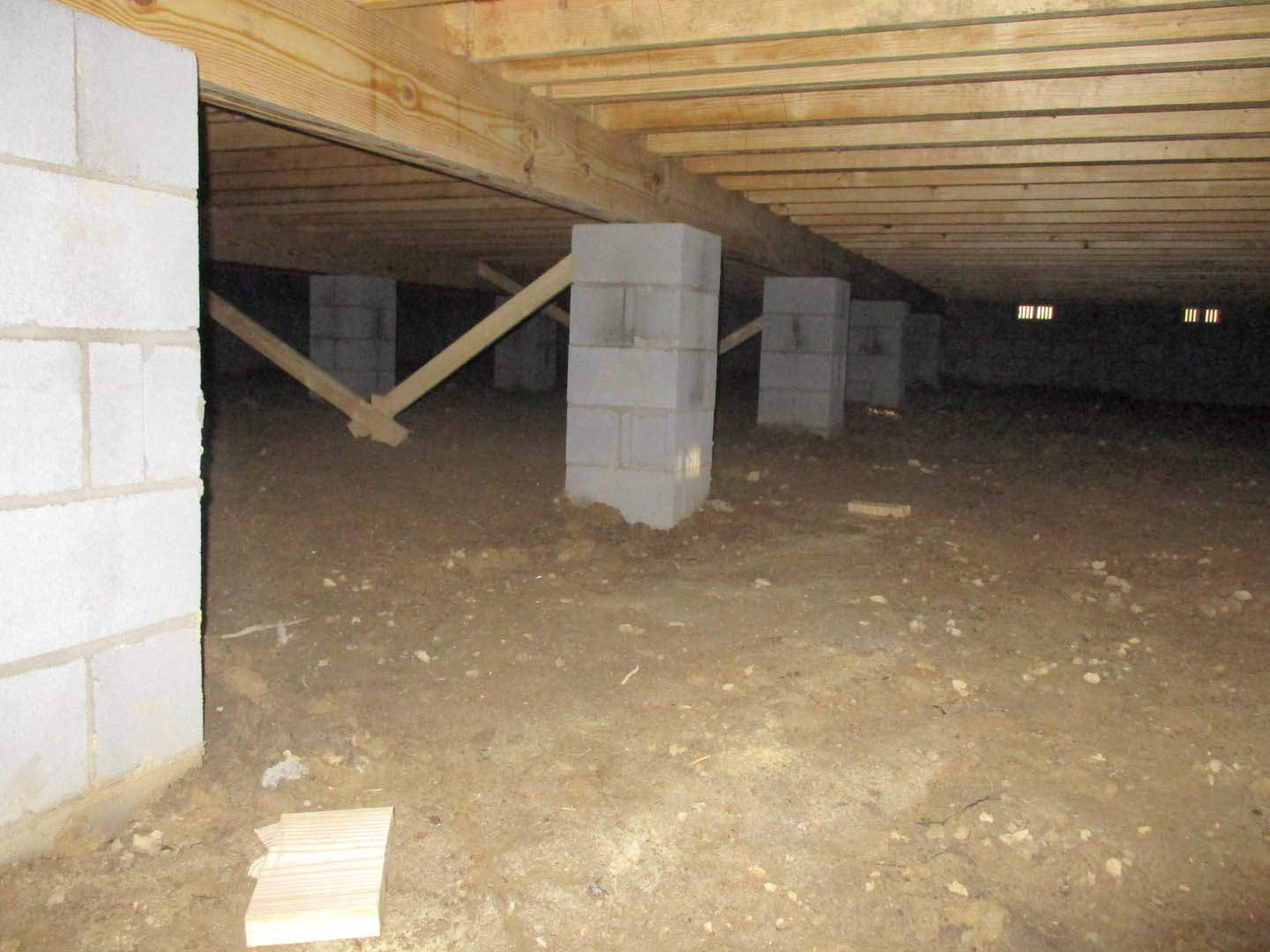 White concrete block basement wall with a black hole, concrete pillar, wooden beam under unfinished ceiling, and piece of wood on bare floor