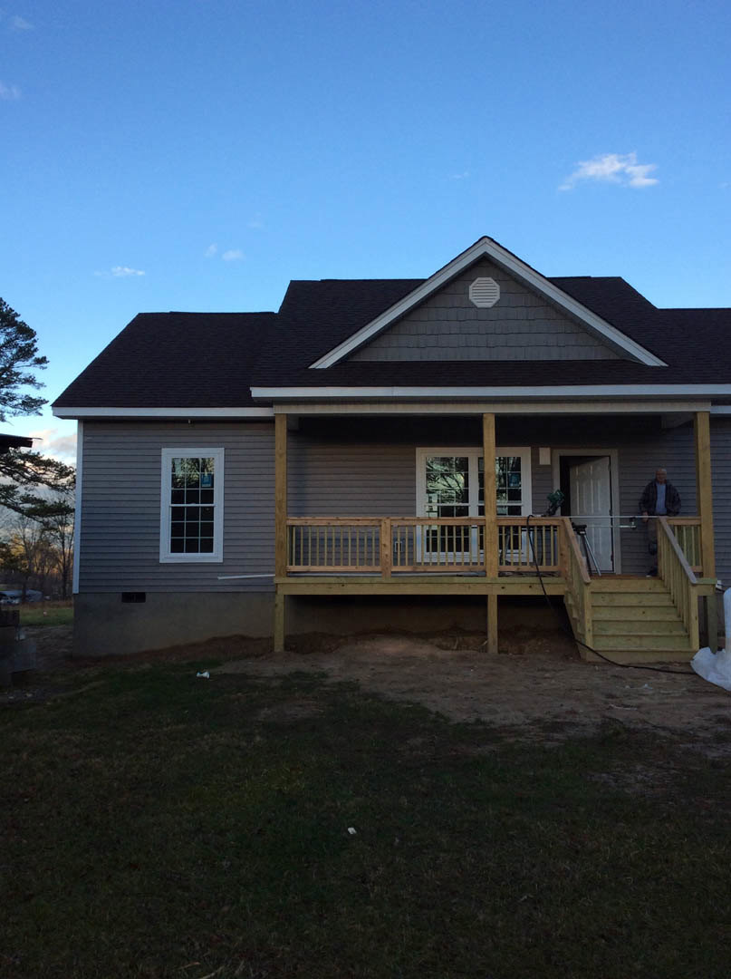 Two-story home with white siding, multi-pane windows, wooden porch and railing, man standing on porch, grassy lawn in foreground, cloudy sky above