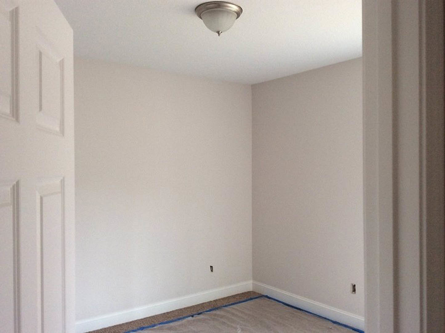 White ceiling-mounted light fixture with a round white shade, white plaster walls, white door with silver handle, beige carpet flooring, and decorative crown molding.