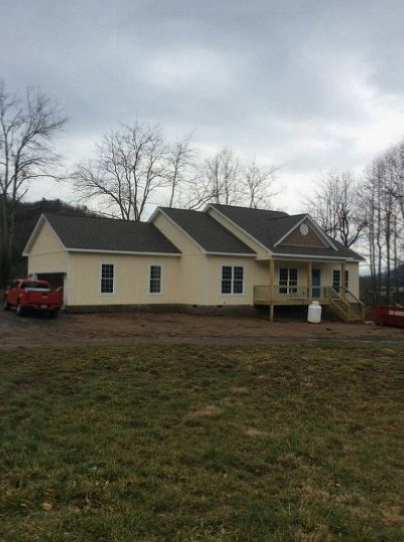 Two-story house with black roof, white-framed windows, and beige siding, red pickup truck parked on gravel driveway, grassy yard bordered by trees, partly cloudy sky overhead