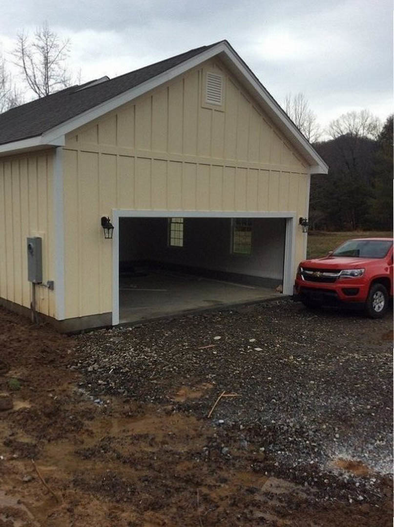 Red pickup truck parked in front of a white garage, muddy driveway with scattered rocks, house roof visible behind, trees and cloudy sky in background.
