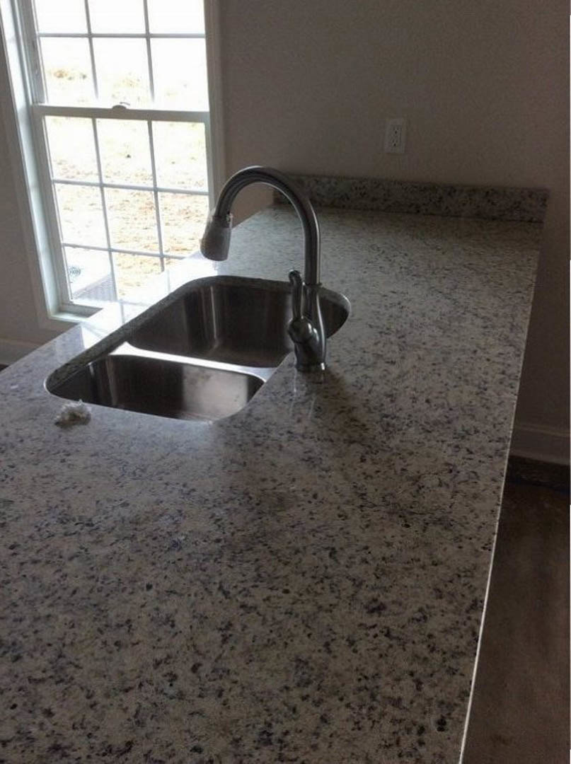 Matte black kitchen sink with stainless steel faucet set into light stone countertop, white tile backsplash visible in background