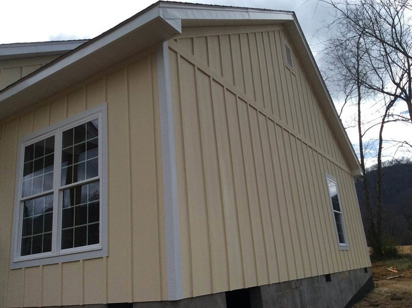White exterior siding with a corner window featuring a white frame, tree branches partially visible in foreground, sloped roof and blue sky in background