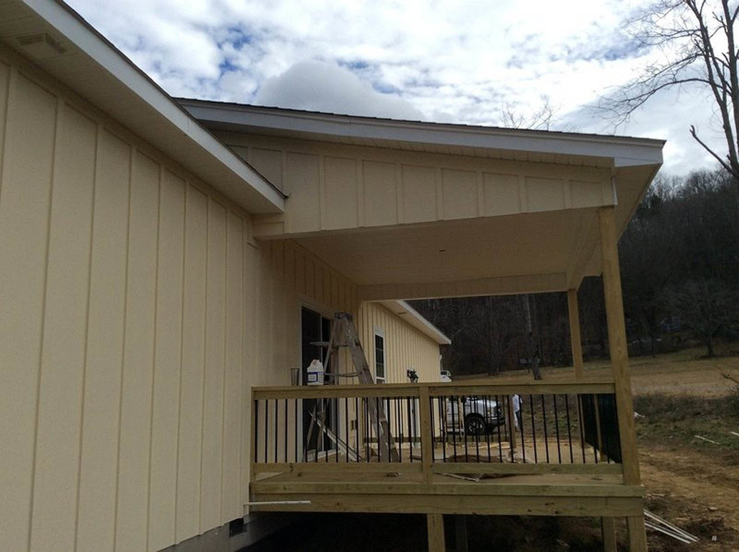 Wood-sided house with fenced deck, wooden ladder leaning against white box, car parked in background, cloudy sky overhead, trees and dirt surface visible