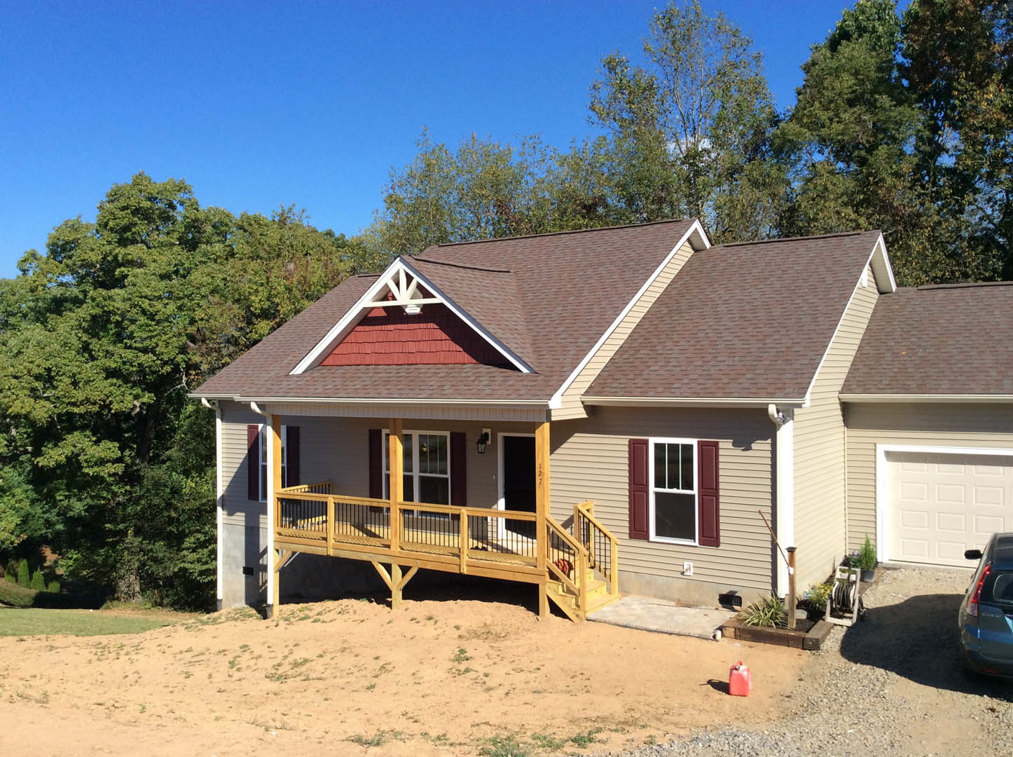 Two-story home with gray siding, wooden deck and railing, stairs leading to backyard, large windows, garage with white door, car parked in driveway, mature trees in background
