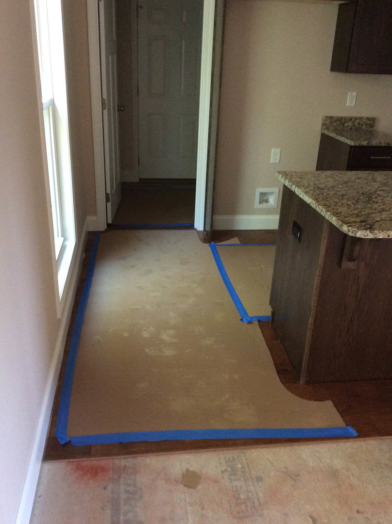 Blue painter’s tape marking edges of light-colored tile flooring near white door with silver handle, plywood subfloor, and adjacent cabinetry.